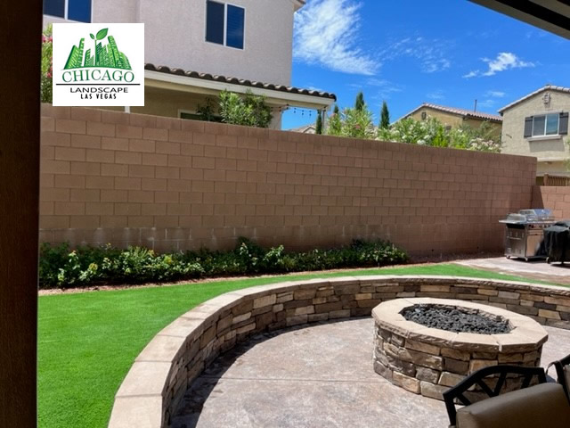 Custom circular gas fire pit inside a curved decorative paver wall beside artificial turf.